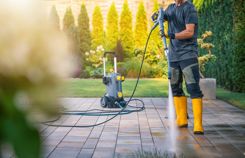 What Sets DewDrop's Pressure Washing Services Apart? A man in yellow boots and gloves operates a pressure washer, cleaning a stone patio surrounded by lush greenery. The garden is sunny, showcasing vibrant plants and a well-maintained outdoor space.