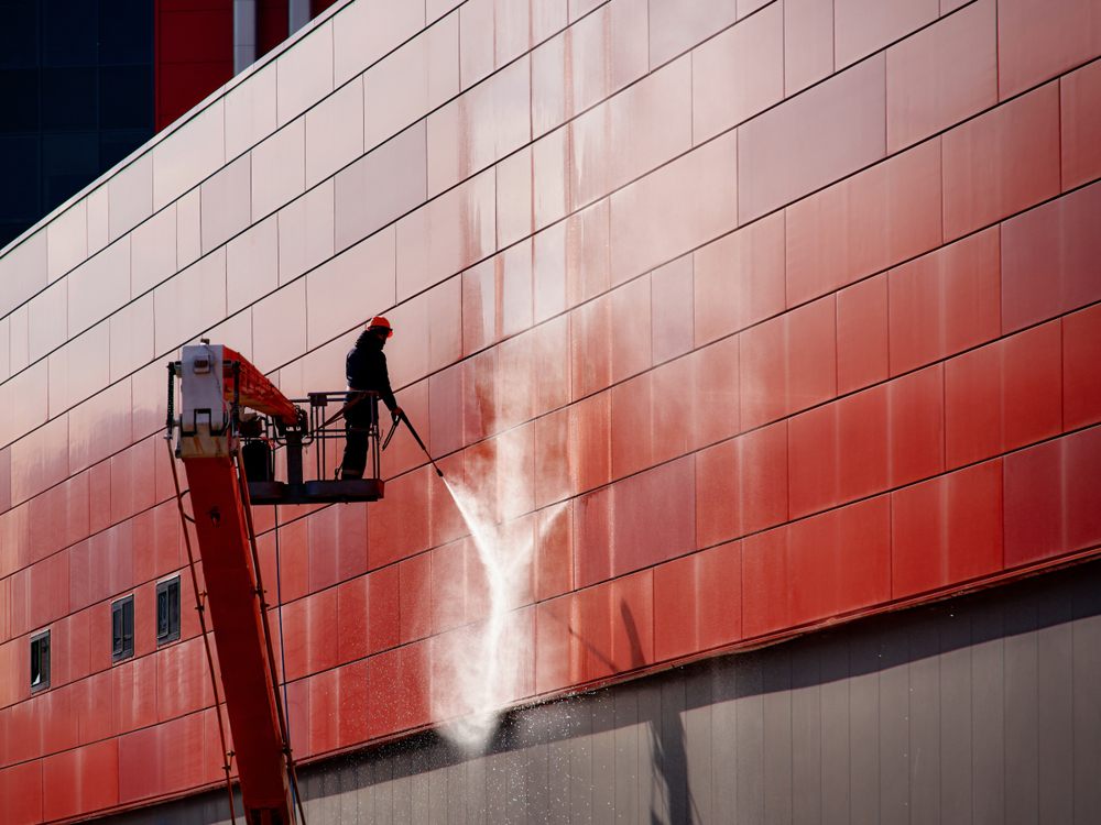 How Pressure Washing Increases Property Value for Residential & Commercial Buildings. Worker wearing safety harness washes wall facade at height on modern building in a crane.