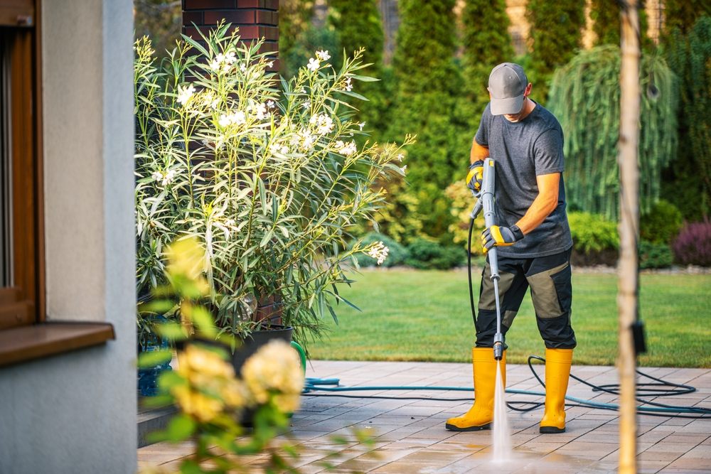 The Environmental Impact of Eco-Friendly Pressure Washing Solutions. A person wearing yellow rubber boots and a gray cap uses a pressure washer to clean a patio in a residential backyard. The sun shines brightly on the scene, highlighting the manicured lawn and vibrant.